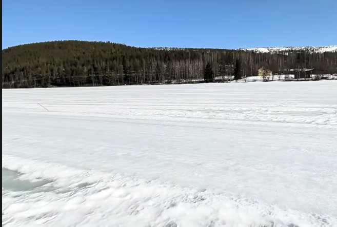 Ampio paesaggio invernale con un lago ghiacciato e una fitta foresta sotto un cielo azzurro. Una piccola casa gialla è visibi