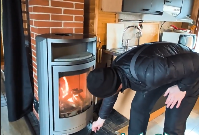 Stefano Mini accende il fuoco in una stufa a legna moderna, in un ambiente rustico con parete in mattoni e angolo cucina.