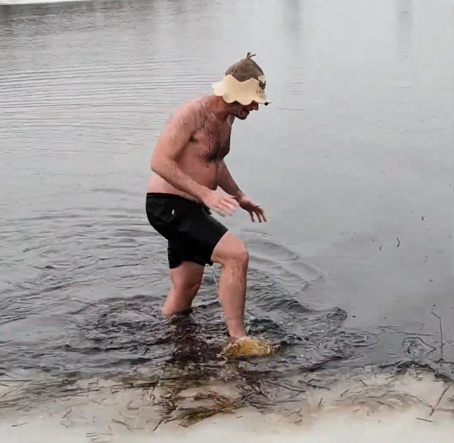 Stefano Mini cammina nell'acqua fredda indossando un cappello da sauna e pantaloncini neri, con detriti sulla riva.