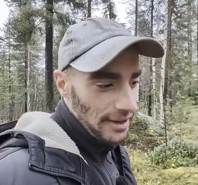 Stefano Mini con cappellino grigio e giacca scura in una foresta, circondato da alberi e vegetazione verde.