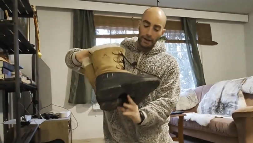 Stefano Mini mostra un paio di stivali invernali beige e neri con lacci in un ambiente interno.