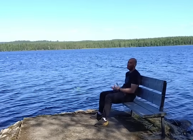 Stefano Mini seduto su una panchina di legno in riva a un lago blu, con una foresta verde sullo sfondo sotto un cielo sereno.