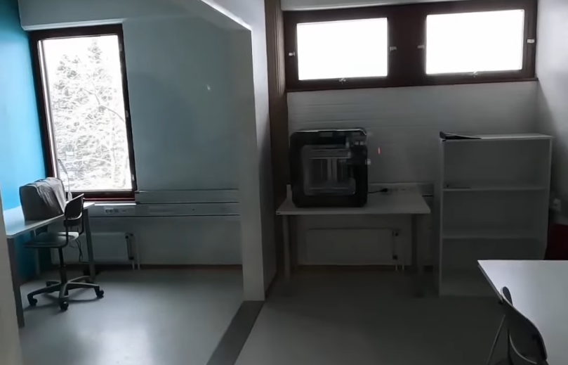 Interno di un ufficio o aula vuota con scrivanie, sedie, finestre e una stampante 3D su un tavolo.
