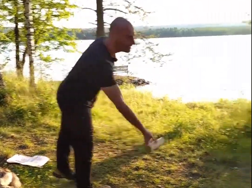 Stefano Mini in maglietta nera e pantaloni scuri, tiene un oggetto cilindrico vicino a un lago con alberi e erba.