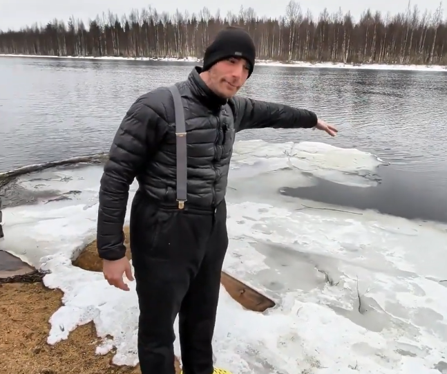 Stefano Mini in piedi su una riva innevata e ghiacciata, indica il fiume con blocchi di ghiaccio galleggianti. Sfondo di fore