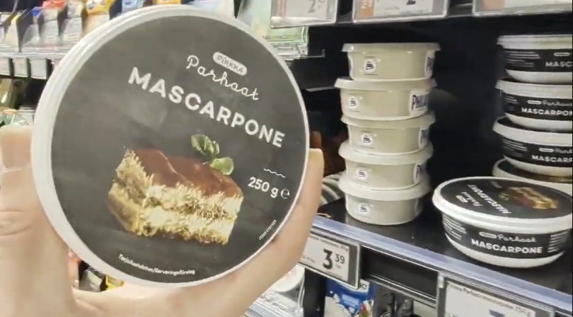 Mano che tiene una confezione di Mascarpone Pirkka Parhaat con immagine di tiramisù, in un corridoio di supermercato.