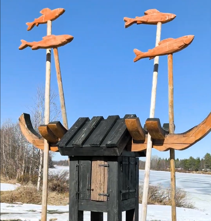 Scultura in legno con pesci e una piccola capanna, su un paesaggio innevato con lago ghiacciato e cielo blu.