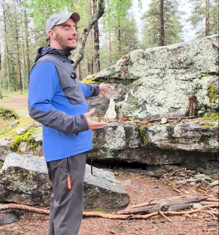 Stefano Mini in un bosco roccioso, indossa una giacca blu e grigia, gesticola mentre presenta un sito storico o naturale.