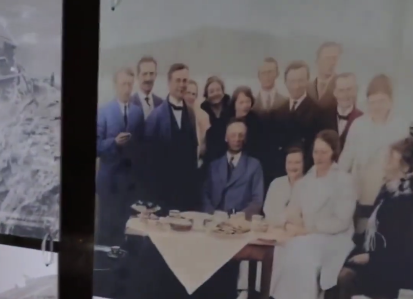 Foto d'epoca di un gruppo di uomini e donne riuniti attorno a un tavolo con cibo e bevande.