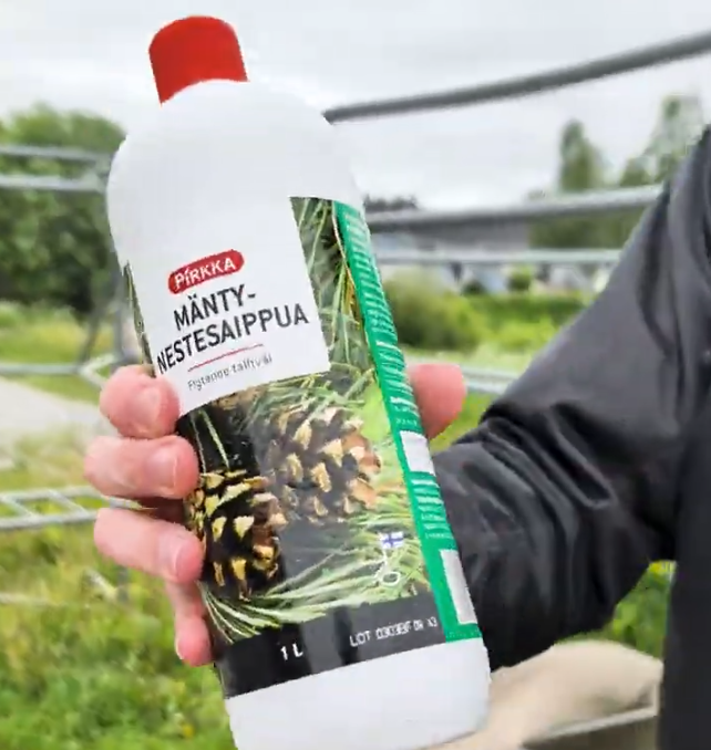 Mano di Stefano Mini che tiene una bottiglia di sapone liquido al pino Pirkka con etichetta verde e rossa, sullo sfondo sfoca