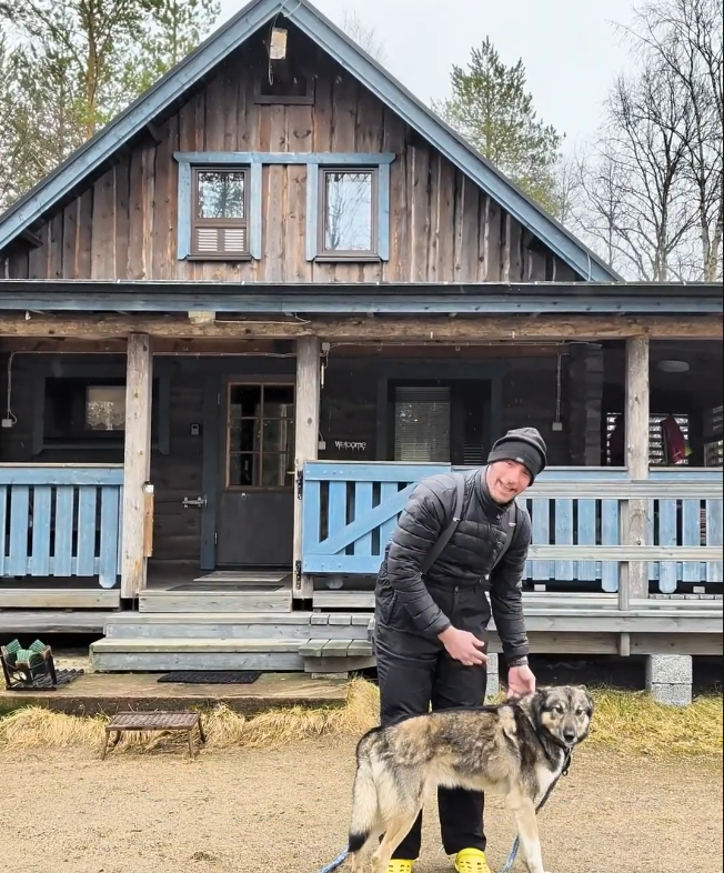 Stefano Mini accarezza un cane husky davanti a una baita di legno con dettagli blu.