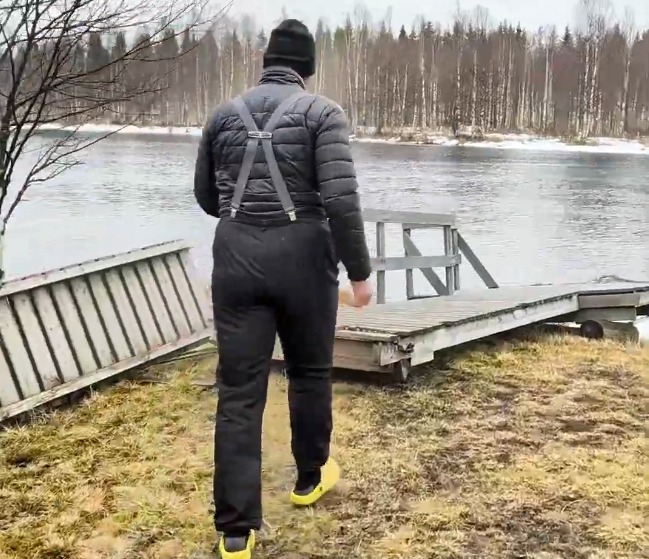Stefano Mini cammina verso un molo di legno su un fiume o lago, indossando abiti invernali e scarpe gialle, con alberi sullo