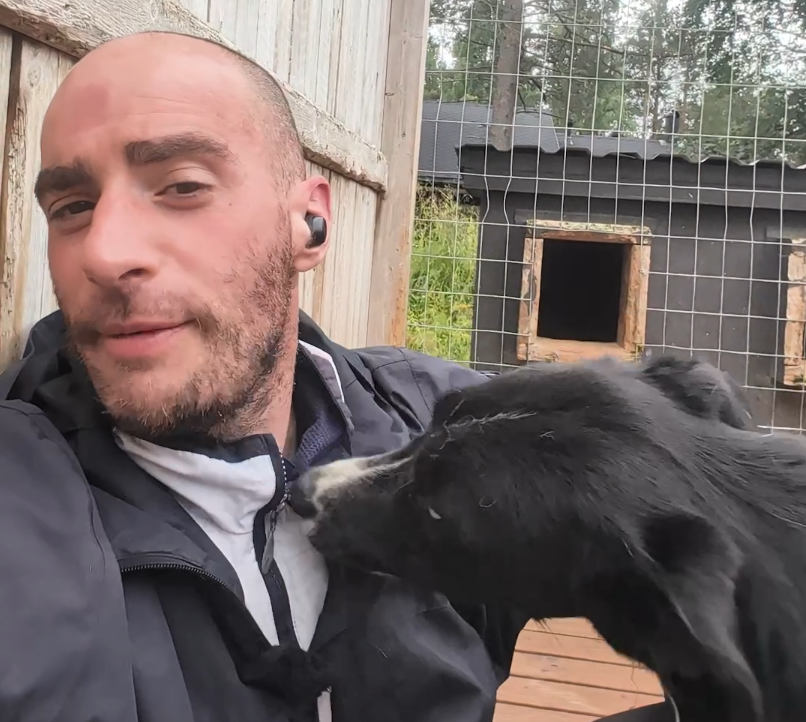 Stefano Mini con un cane nero e bianco che gli lecca il petto, in un ambiente esterno con una cuccia per cani.
