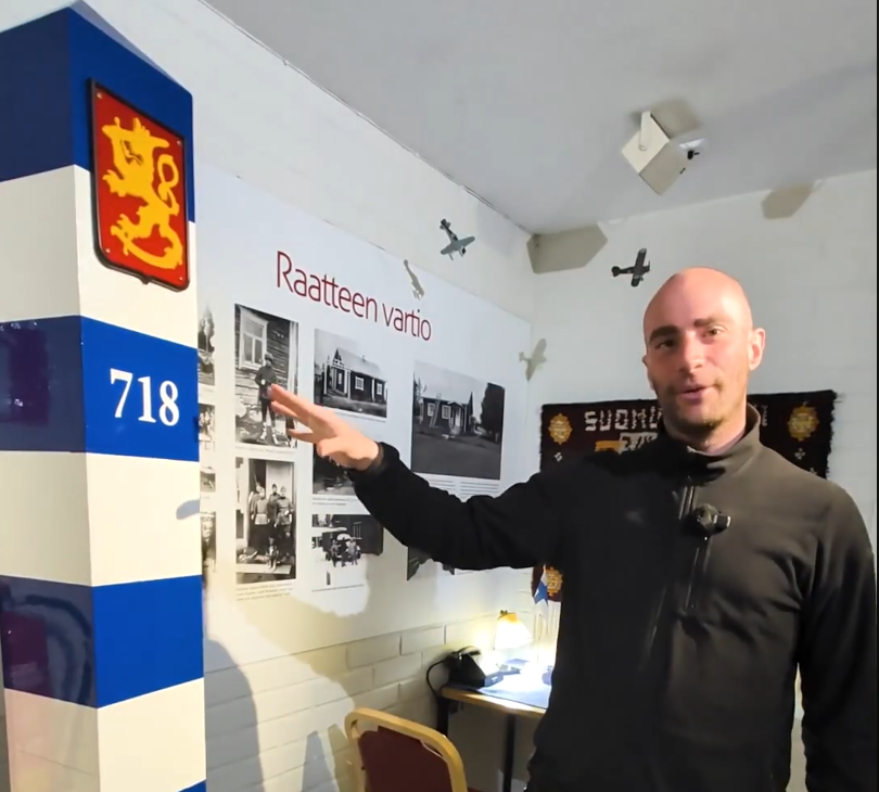 Stefano Mini in un museo finlandese indica una mostra sulla "Raatteen vartio" con foto storiche e un pilastro con lo stemma.