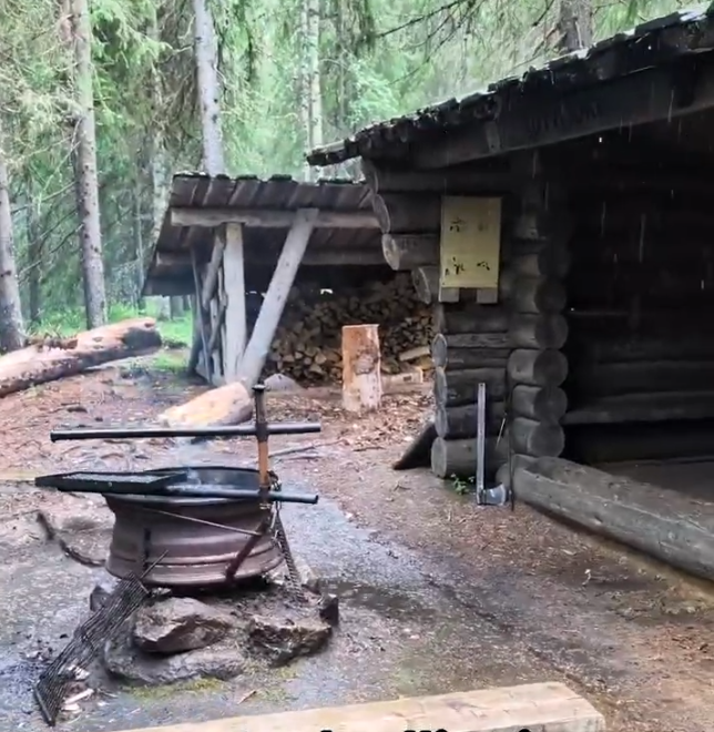 Capanna di legno rustica in una foresta con legna da ardere e un'area per cucinare all'aperto sotto la pioggia.