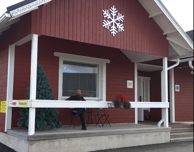 Stefano Mini seduto sulla veranda di una casa rossa con un albero di Natale e un fiocco di neve decorativo.