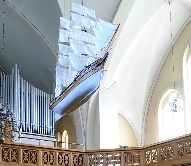 Modello di veliero a vele spiegate sospeso al soffitto di una chiesa, con organo a canne e balcone in legno intagliato.