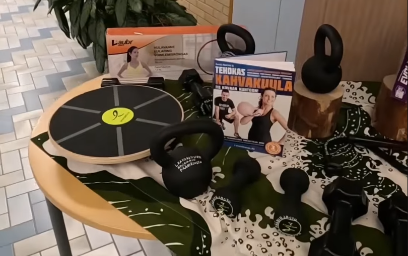 Assortimento di attrezzature fitness su un tavolo, inclusi kettlebell, manubri, balance board e il libro 'Tehokas Kahvakuula'