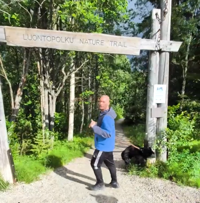 Stefano Mini cammina su un sentiero naturale nel bosco, sotto un cartello che indica 'Nature Trail', con un cane nero al suo