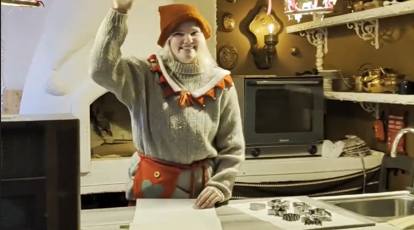 Donna sorridente vestita da elfo con cappello arancione e grembiule rosso, saluta in una cucina rustica con formine per bisco