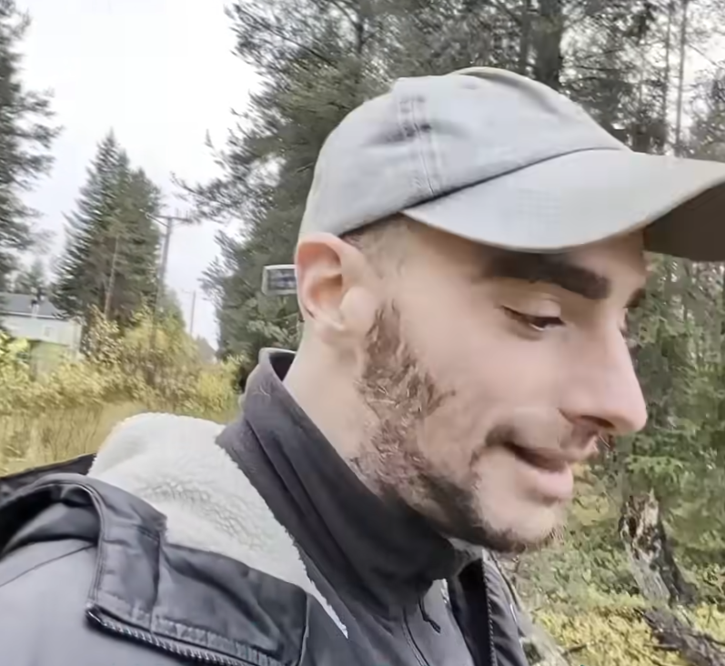 Stefano Mini, con cappellino e barba, indossa una giacca in un ambiente naturale con alberi e vegetazione sullo sfondo.