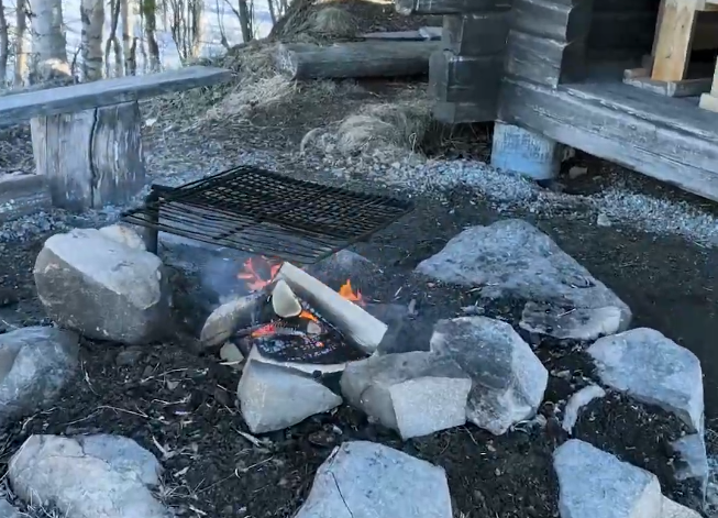 Falò con griglia per barbecue in un'area picnic rustica, circondato da rocce e una panca di legno.