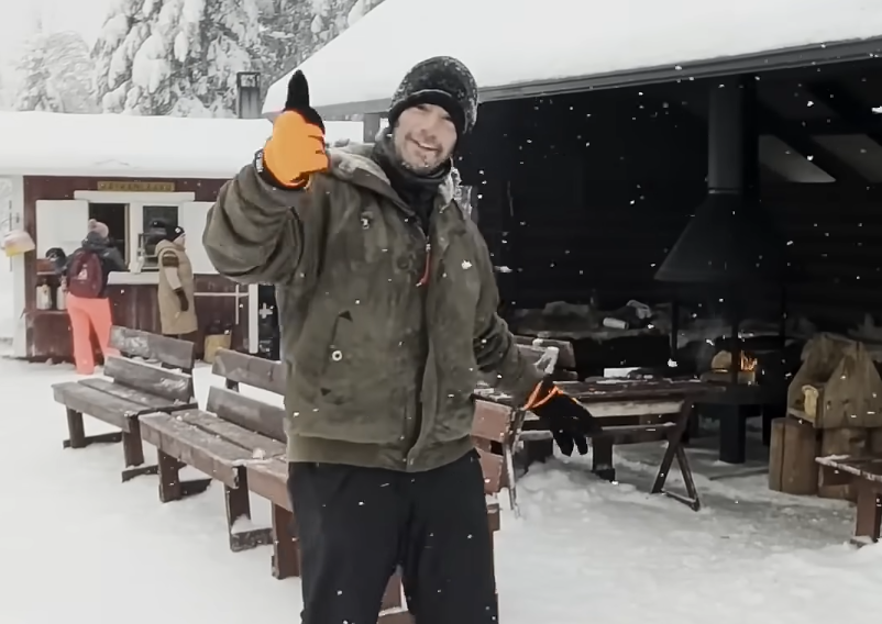 Stefano Mini sorridente con pollice in su in un paesaggio innevato, con baite e persone sullo sfondo.