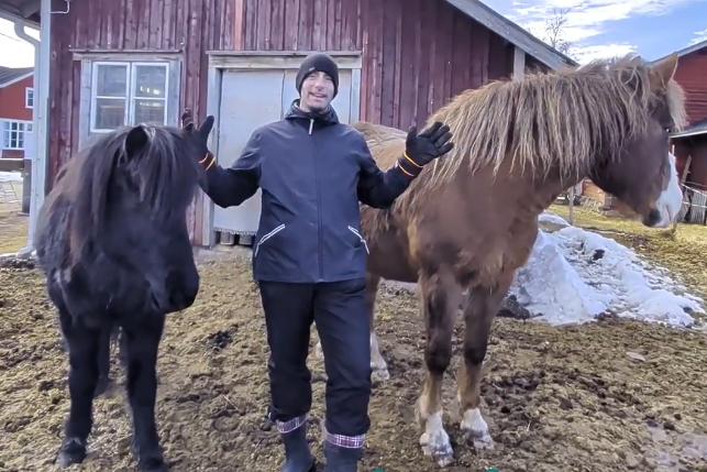 Stefano Mini tra un pony nero e un cavallo marrone, all'aperto con neve e un edificio rosso sullo sfondo.
