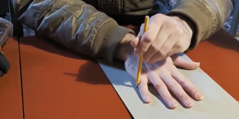 Una mano traccia il contorno di un'altra mano su un foglio di carta con una matita, su una superficie marrone-rossastra.