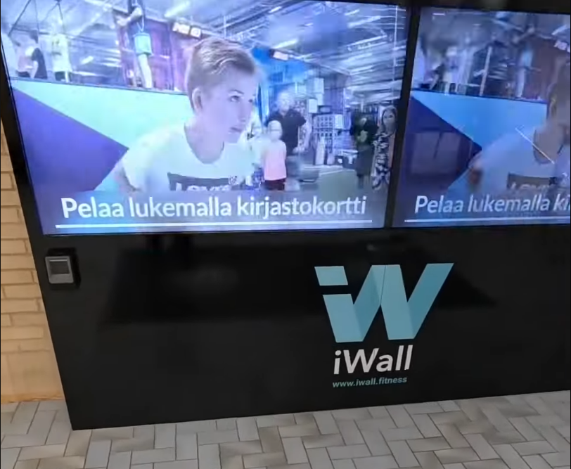 Display interattivo iWall con una persona che gioca a un videogioco, con il testo "Pelaa lukemalla kirjastokortti".