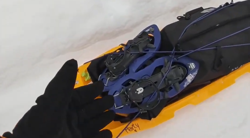 Mano guantata che indica una slitta gialla con racchette da neve blu e una borsa nera sulla neve.