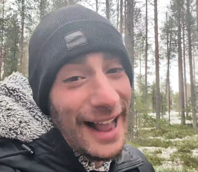 Stefano Mini sorride in un bosco innevato, indossando un berretto nero e una giacca calda.