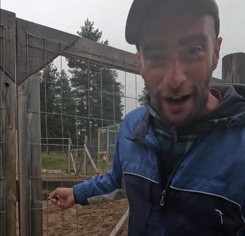 Stefano Mini in giacca blu e cappello, in piedi davanti a un recinto di rete metallica in un'area esterna con alberi.