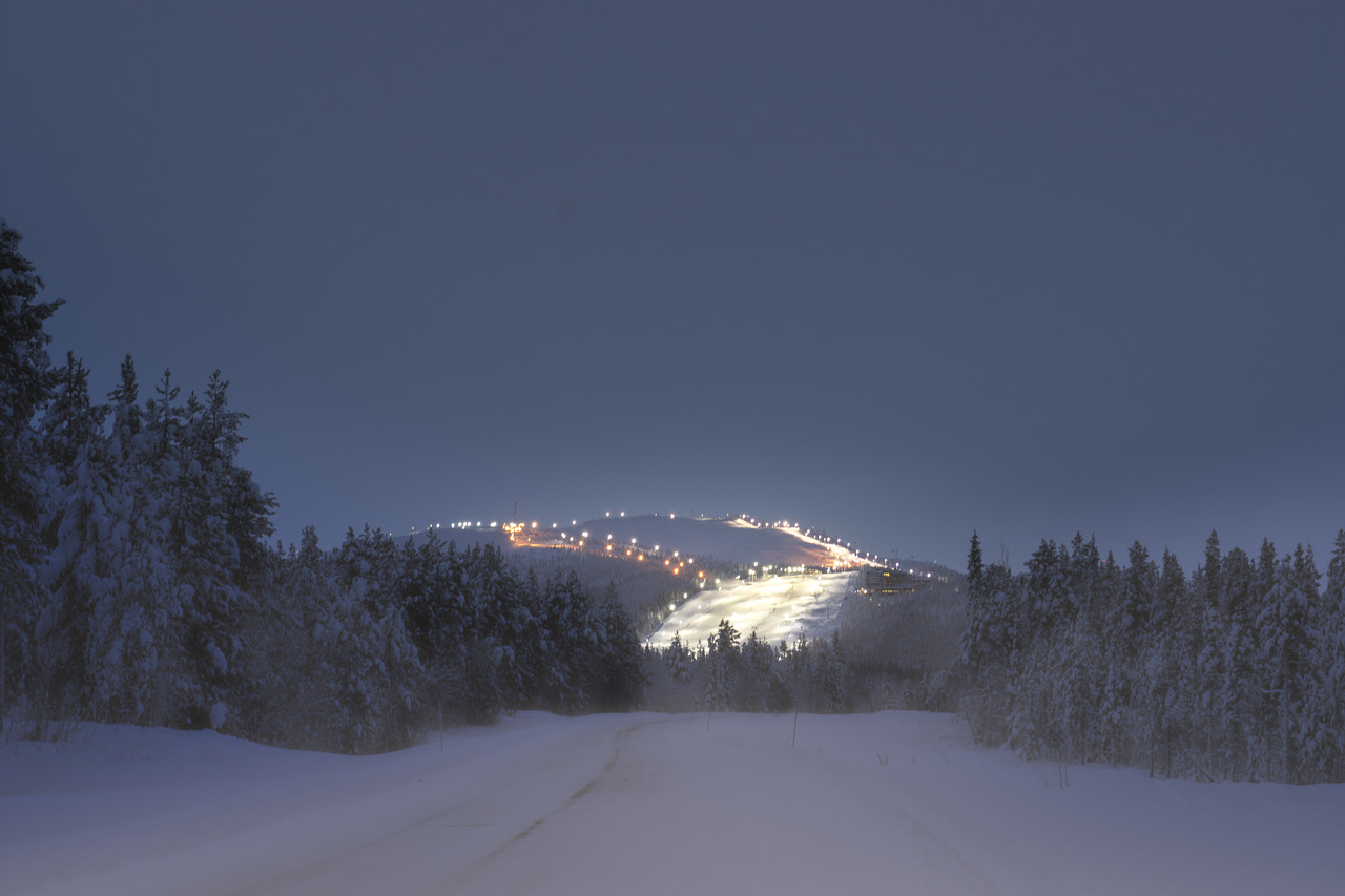 Paesaggio invernale notturno con strada innevata, alberi coperti di neve e pista da sci illuminata in lontananza.