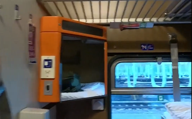 Interno di un vagone letto di un treno con una cuccetta arancione e un finestrino che mostra un paesaggio invernale innevato.