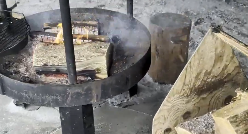 Focolare in metallo con legna che brucia, fumo che sale e una griglia. Accanto, ceppi di legno per il fuoco.