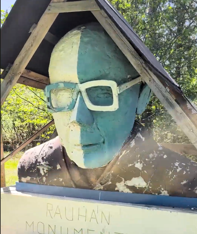 Busto di una scultura con occhiali, dipinta a metà in bianco e a metà in verde acqua, sotto una tettoia di legno, parte del M
