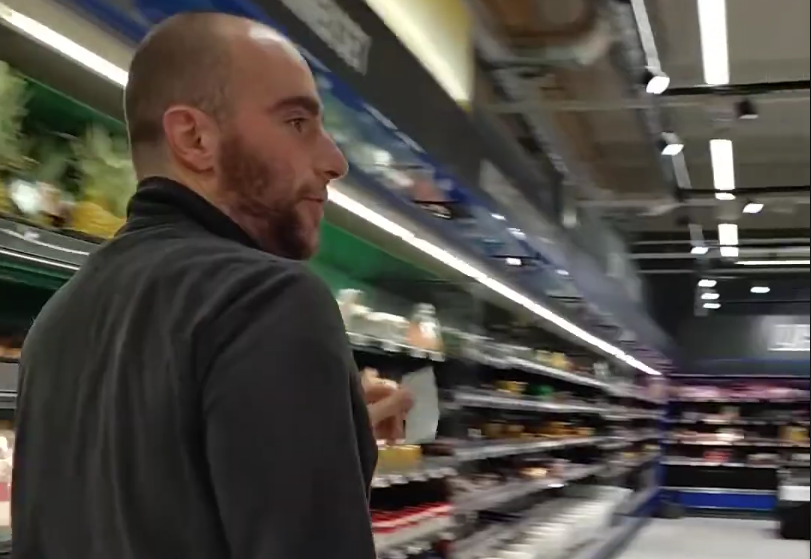 Stefano Mini cammina lungo un corridoio di supermercato, con scaffali pieni di prodotti e luci brillanti sopra.