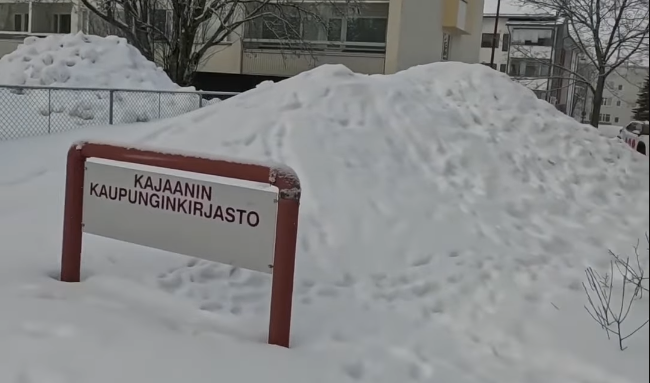 Segnale della Biblioteca Civica di Kajaani in un paesaggio innevato con grandi cumuli di neve e edifici sullo sfondo.