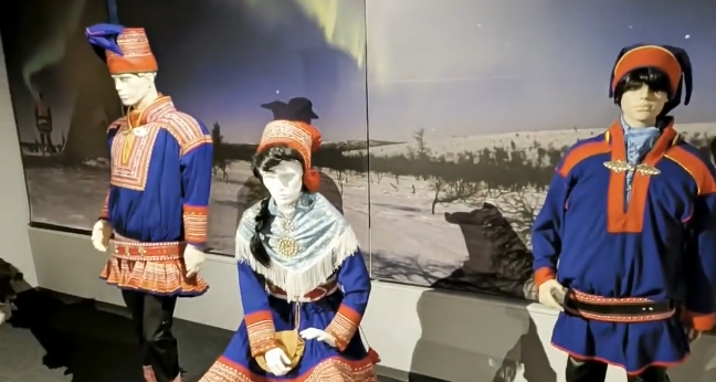 Manichini con abiti tradizionali Sami in un museo, con sfondo di paesaggio invernale e aurora boreale.