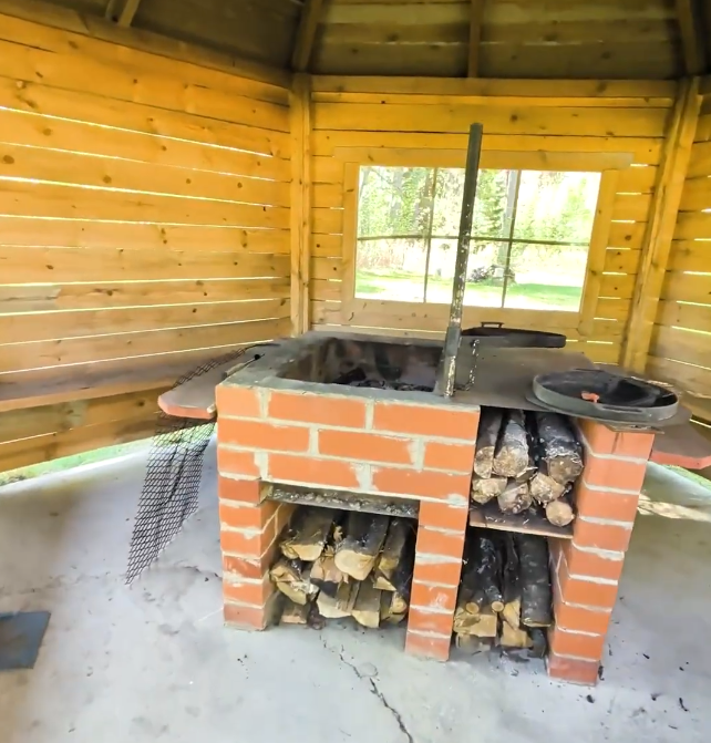 Barbecue in mattoni con legna da ardere in una struttura in legno all'aperto, con finestra che mostra alberi verdi.