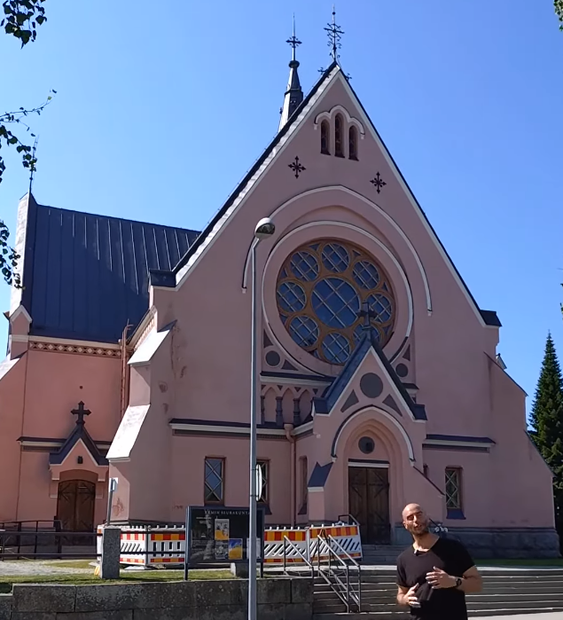 Stefano Mini in piedi davanti a una chiesa rosa con tetto scuro e grande rosone circolare sotto un cielo azzurro.