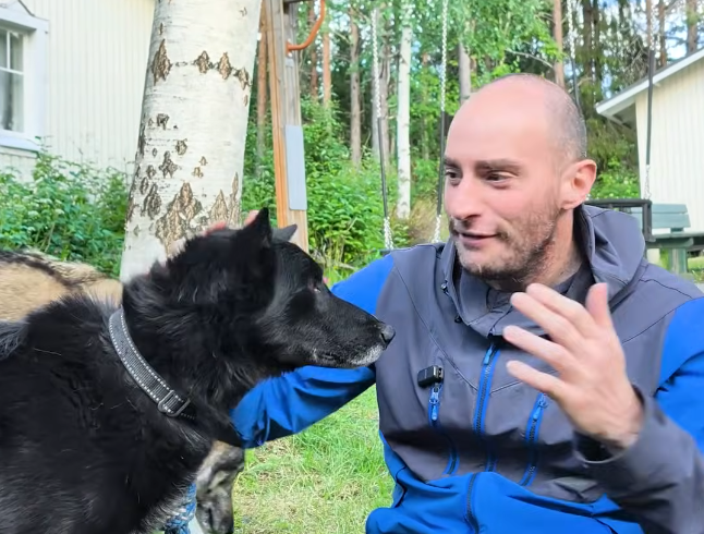 Stefano Mini accarezza un cane nero con collare all'aperto, con alberi e una casa bianca sullo sfondo.