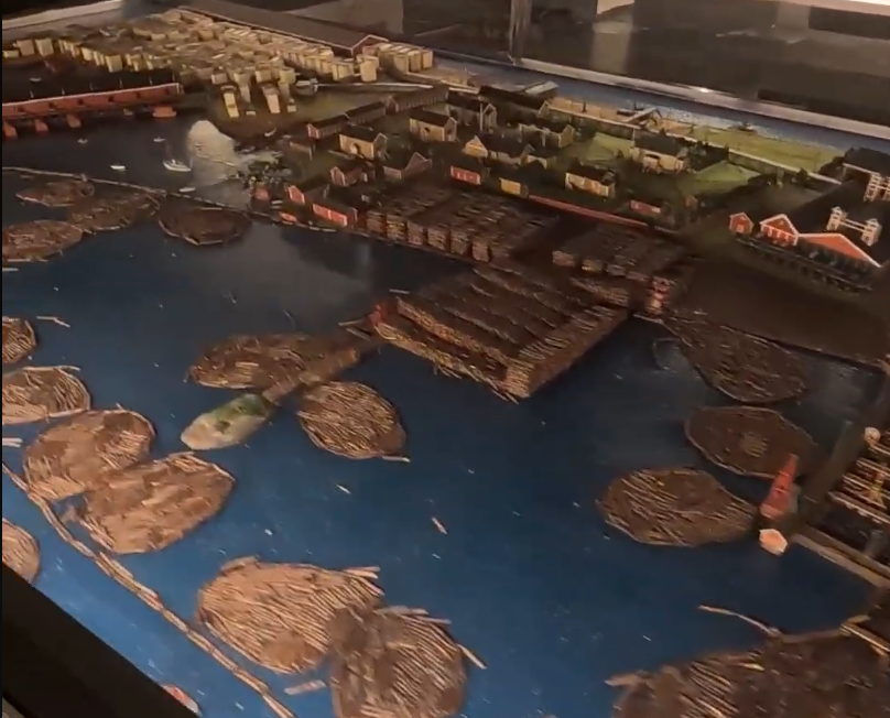 Modello in miniatura dettagliato di un villaggio costiero con edifici, un ponte e tronchi galleggianti nell'acqua.