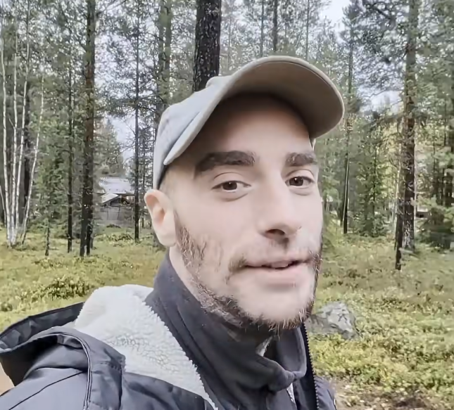 Stefano Mini in un bosco con alberi e vegetazione, indossa un cappellino e una giacca. Sullo sfondo si intravede una baita.