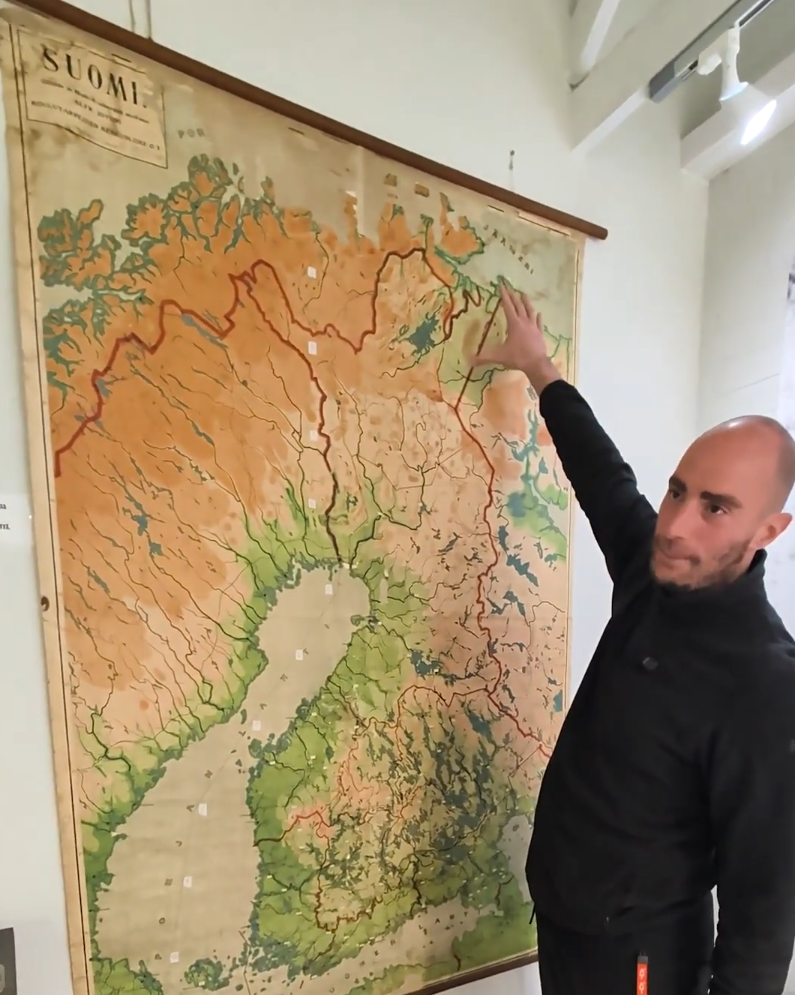 Stefano Mini indica una vecchia mappa della Finlandia appesa a una parete bianca, mostrando dettagli geografici del paese.