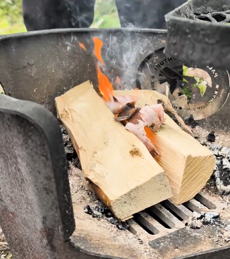 Legna che brucia in un braciere di metallo, con fiamme vivaci e fumo che sale, creando brace per cucinare.