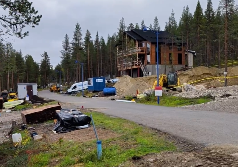 Cantiere edile in un'area boschiva con una casa in legno in costruzione, cumuli di terra, un escavatore e materiali edili.