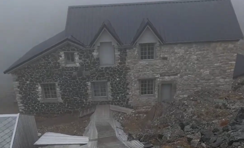 Antico edificio in pietra con tetto scuro e finestre a griglia, immerso nella nebbia su un terreno roccioso.