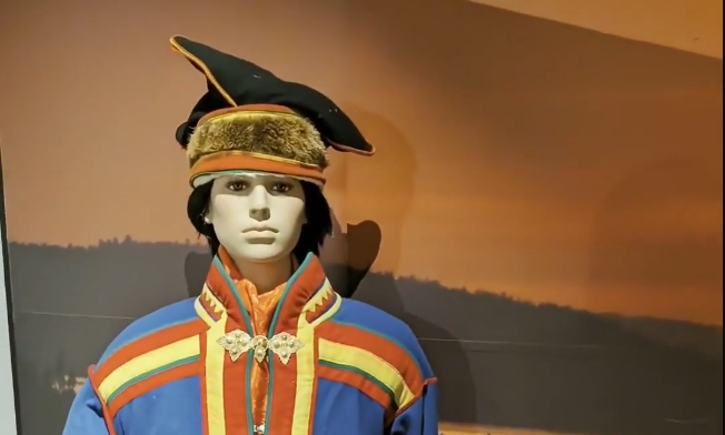 Manichino con abito tradizionale Sami blu, rosso e giallo, con cappello nero e pelliccia, su sfondo paesaggio al tramonto.